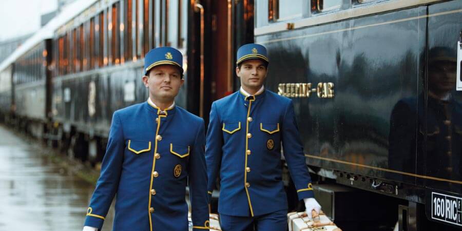 Two members of Venice Simplon-Orient-Express staff, dressed in their iconic blue uniform, carry luggage on the train platform. 